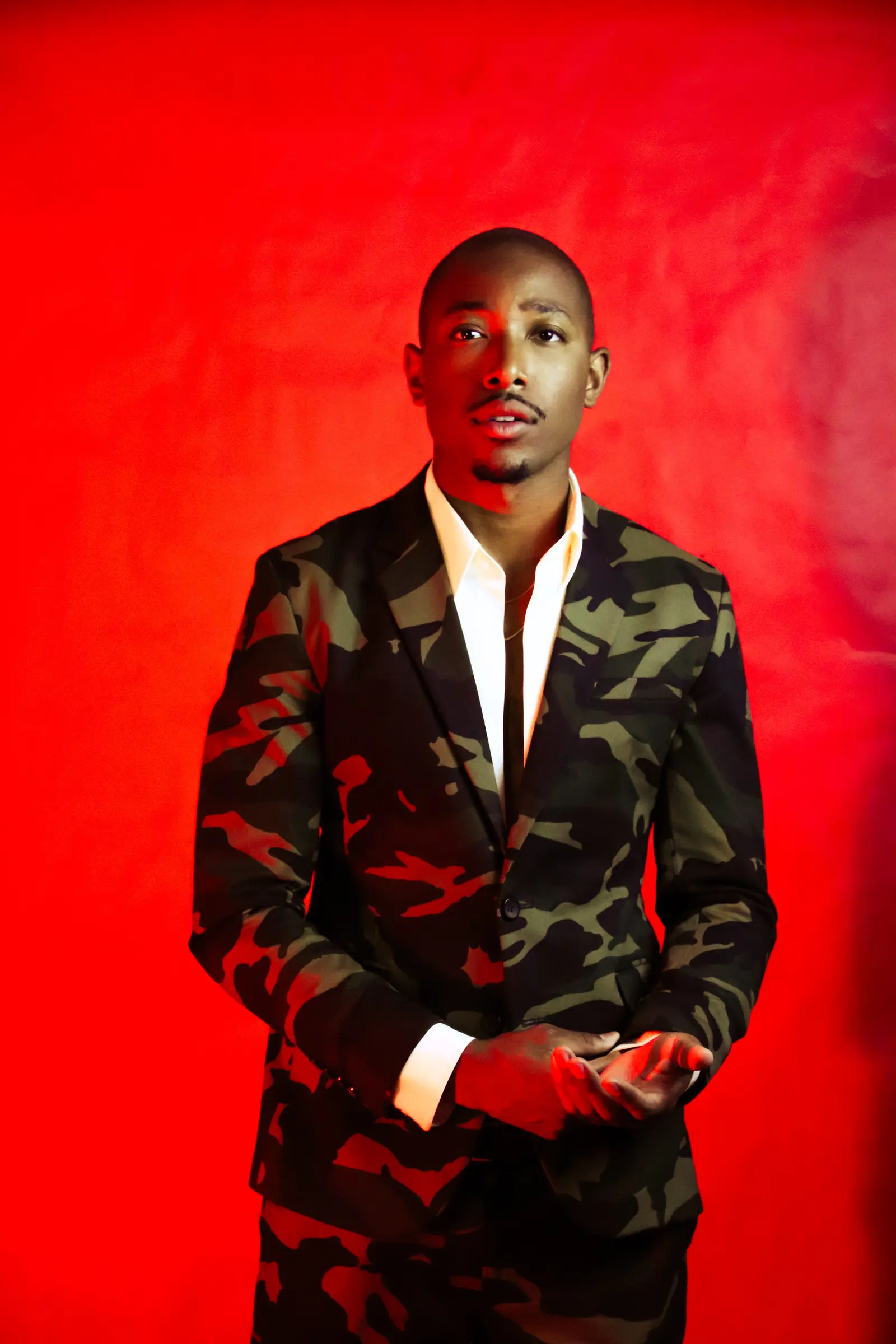 Full-length portrait of Malcolm Xavier in a camouflage suit against a red studio backdrop.